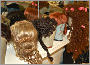 image of four wigs, 1 blond with a bun, one brown with a braid and curls, one short bright red and one dark red with curls and flowers, on a counter reflected in a mirror. 